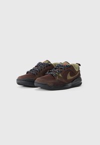 Brune sneakers laget av mesh og semsket skinn, med svarte gummisåler, grønne detaljer og mønstrede svarte og hvite snører. Fremtredende merkevare tilstede.