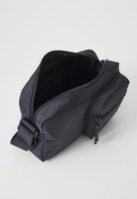 Borsa a tracolla in tessuto nero con tracolla regolabile, scomparto principale con zip e piccola tasca frontale con dettaglio zip.