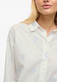 Camicia bianca con bottoni, caratterizzata da ricami floreali blu chiaro, colletto classico e vestibilità rilassata. Tessuto morbido con una consistenza liscia.
