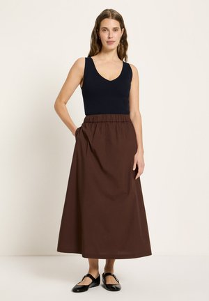 Frau mit langen braunen Haaren, die ein ärmelloses schwarzes Ripptop, einen hoch taillierten braunen Midirock und schwarze Mary Janes trägt, mit einer Hand in der Tasche stehend.