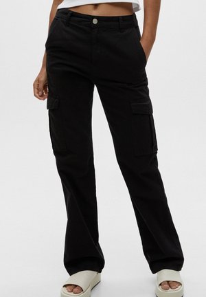Femme portant un pantalon cargo noir avec des poches à rabat et des sandales compensées blanches à bout ouvert, debout devant un fond uni.