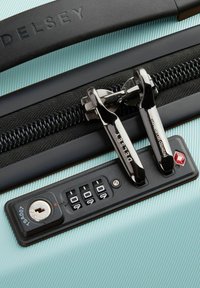 Un zipper de color verde menta en una maleta con tiradores cromados dobles, un candado de seguridad negro y un disco de combinación, con el logo de Delsey grabado.