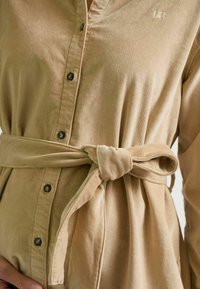 Eine Nahaufnahme eines hellbraunen Cordhemds mit V-Ausschnitt und Knöpfen, das an der Taille mit einem gebundenen Gürtel betont wird, wobei Textur und Stil hervorgehoben werden.