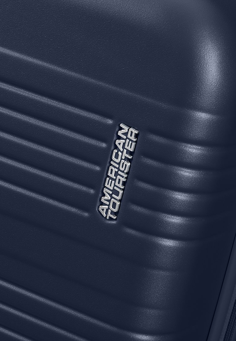 Angolo di una valigia rigida blu navy con scanalature orizzontali in rilievo e logo argentato "American Tourister" dettagliato sulla superficie.