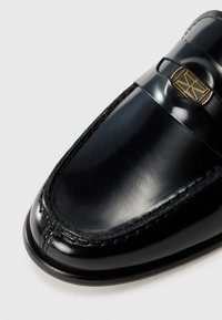Mocassini in pelle nera con un design elegante, superficie lucida, cucitura rialzata lungo i bordi e un accento in tono oro sulla banda.