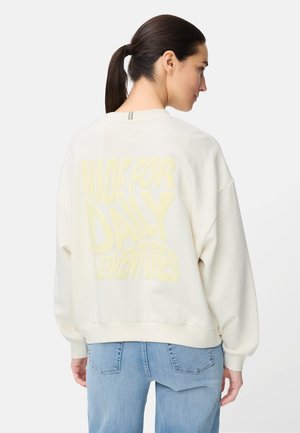 Femme aux cheveux foncés attachés en queue de cheval, portant un sweat-shirt blanc ample avec un texte jaune "Made for Daily Adventures" et un jean bleu clair, vue de dos.
