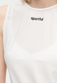 Žena oblečená v bezrukávovom bielom topu s nápisom "sportful" vytlačeným čiernou blízko goliera na hornej časti hrudníka.