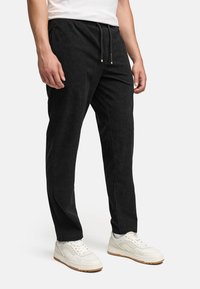 Mies, joka käyttää mustia corduroy-housuja, joissa on nauhavyötärö ja valkoiset tennarit, seisoo yksinkertaista valkoista taustaa vasten.