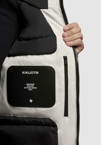 Polstret jakke indvendigt, der viser en sort lomme med hvid tekst "KHUJOTM," "GENUINE BRAND," "ESTABLISHED 2001," og et lille ahornblad.