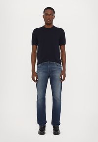 Marineblauwe t-shirt met korte mouwen, lichtblauwe straight-leg jeans en zwarte nette schoenen. Model staat tegen een neutrale achtergrond.