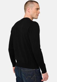 Mann in schwarzem Langarmpullover und blauen Jeans, seitlich gedreht, zeigt Rücken und Profil vor hellem einfarbigem Hintergrund.