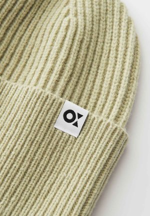 Hellbeige ribbed Strickmütze mit umgeschlagenem Rand und einem weißen rechteckigen Etikett mit einem schwarzen geometrischen Logo.