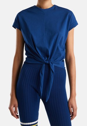 Mujer con una blusa azul de manga corta anudada al frente y pantalones azules acanalados de talle alto con rayas blancas y amarillas cerca de las rodillas.