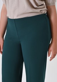 Persoon in smaragdgroene high-waisted broek en een beige top met korte mouwen en kleine sprankelende details, tegen een effen licht achtergrond.