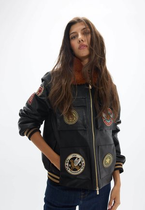 Joven mujer con cabello largo y castaño que lleva una chaqueta bomber negra con parches y un cuello de piel marrón, de pie con una mano en el bolsillo.