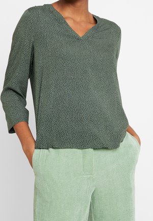 Kobieta w ciemnozielonej bluzce z długim rękawem w białe kropki oraz jasnych zielonych prążkowanych spodniach, z rękami w kieszeniach, na białym tle.