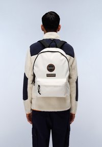 Zaino bianco in canvas, con una tasca frontale, bordi neri e un logo circolare con testo. Indossato su una giacca in pile.