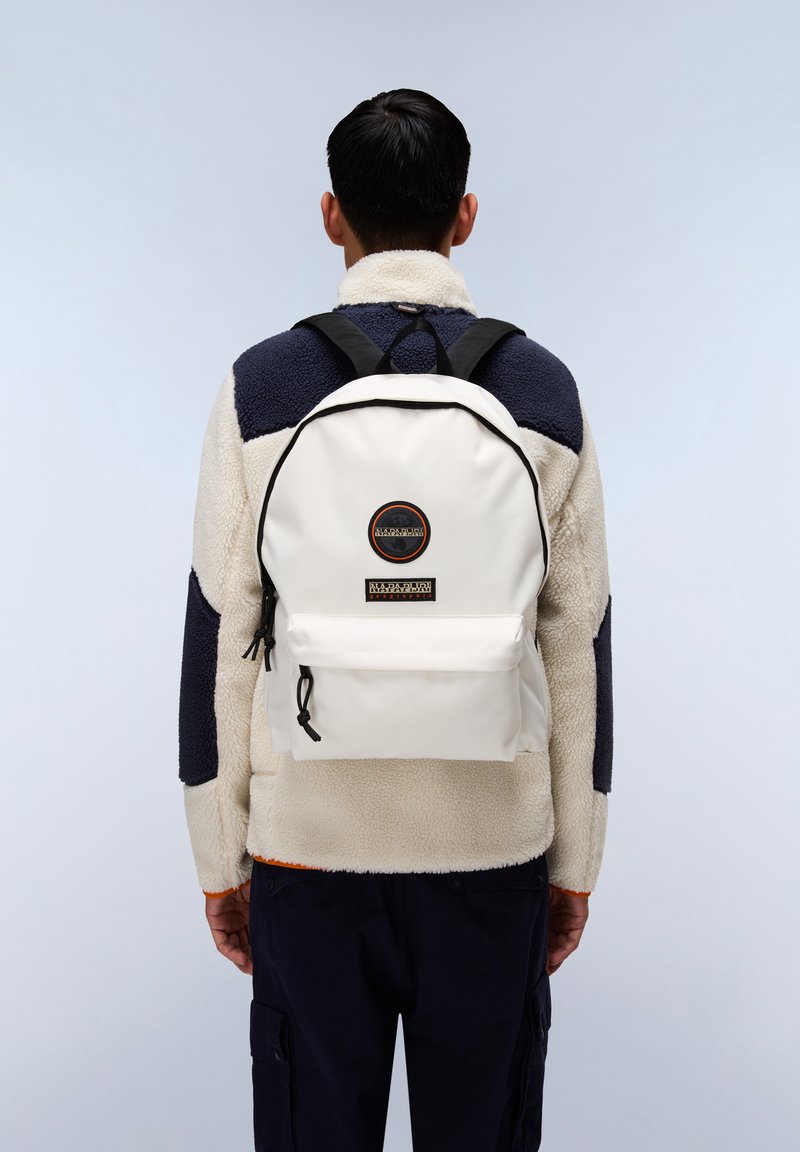 Zaino bianco in canvas, con una tasca frontale, bordi neri e un logo circolare con testo. Indossato su una giacca in pile.