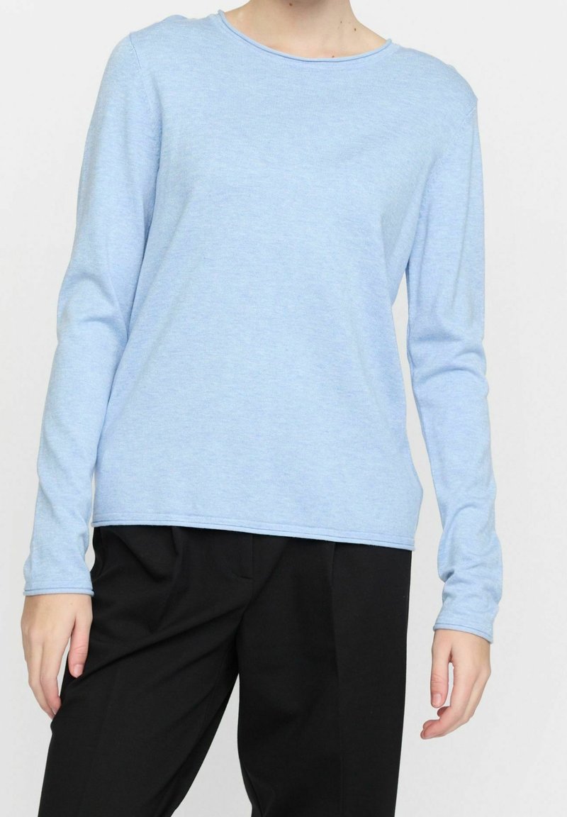 Personne portant une chemise à manches longues bleu clair et un pantalon noir ample, debout devant un fond uni.
