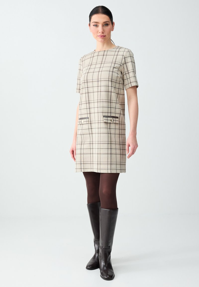 Femme portant une robe à manches courtes à carreaux beige, des collants foncés et des bottes noires hautes jusqu'aux genoux, debout devant un fond clair uni.