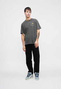 Graues Baumwoll-T-Shirt mit kurzen Ärmeln und einem Frontdruck, kombiniert mit schwarzen Cordhosen und grauen Turnschuhen mit blauen Schnürsenkeln.