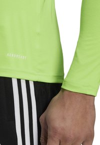 Groene sportshirt met lange mouwen en ribbelstructuur, met het "AEROREADY" logo. Zwarte shorts met drie zichtbare witte strepen.