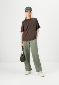Bruine katoenen t-shirt met korte mouwen, gecombineerd met olijfgroene wijde broek, witte sneakers en een donkergroene pet. Klein logo detail op het t-shirt.