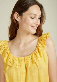 Haut en coton jaune avec un col en dentelle à volants fleuris, doté d'une fermeture à boutons et d'une texture lisse. Motif brodé subtil sur le tissu.