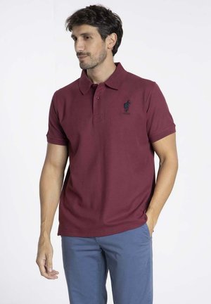 Polo en maron fait de coton, avec un col classique, trois boutons, et un logo brodé bleu foncé sur la poitrine gauche.
