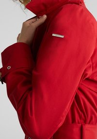 Chaqueta roja con un alto cuello, un acento de logotipo metálico brillante y una cintura con cinturón. Textura suave con detalles de botones en los puños.