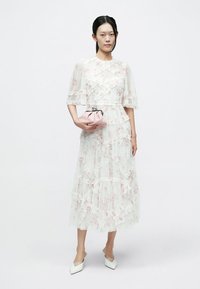 Biała sukienka w kwiatowy wzór z warstwowymi falbanami, krótkie rękawy; modelka trzyma różową kopertówkę i ma na sobie białe buty z noskiem w szpic.