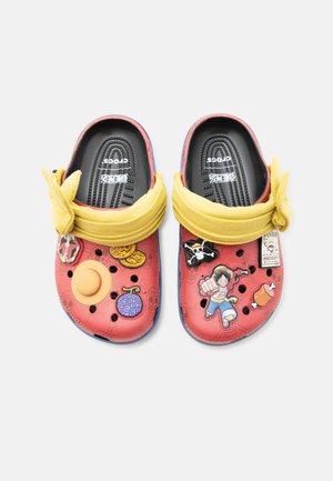 Crocs rouges décorés avec des breloques à l'effigie des personnages de l'anime One Piece, une lanière jaune en tissu nouée en noeud et des semelles intérieures noires texturées.