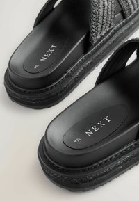 Sandales à plateforme noires avec des sangles tissées, semelle texturée et marquage "NEXT". Le design présente une base épaisse en caoutchouc noir et une finition lisse.