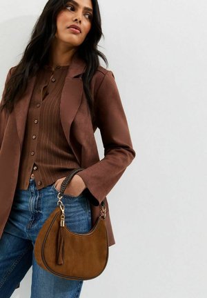 Woman wearing brown suede jacket and ribbed button-up sweater, holding tan suede handbag with braided handle and tassel, paired with blue jeans.