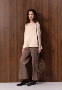 Femme debout contre un mur en bois, portant un haut beige à manches longues, un pantalon large marron, des bottines noires, tenant un sac à main marron.