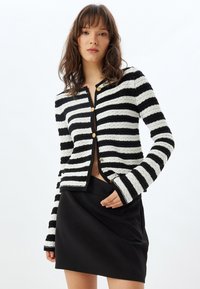 Cárdigan de rayas en blanco y negro con tejido texturizado, cuello redondo y botones dorados. Combinado con una falda mini negra.