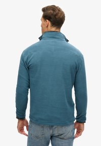 Hombre vestido con una chaqueta de forro polar azul y vaqueros azul claro, de pie con la espalda hacia la cámara sobre un fondo blanco.