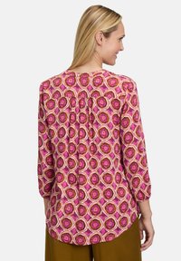 Blusa a maniche lunghe di colore rosa con un motivo geometrico. Presenta un'orlo arrotondato e delle pieghe nella parte posteriore. Realizzata in tessuto leggero.