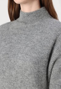 Grauer Strickpullover mit hohem Rippkragen, der eine weiche Textur und einen leicht lockeren Schnitt aufweist. Der Stoff zeigt ein dezentes Muster.