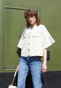 Jaqueta cropped em mistura de algodão branca com dois bolsos frontais, emparelhada com jeans denim azul claro. Modelo segurando uma mala bege, contra um fundo verde.