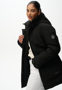 Młoda kobieta w czarnym, pikowanym płaszczu z wysokim kołnierzem, białym swetrze i białych spodniach, pozująca na jednolitym jasnym tle.