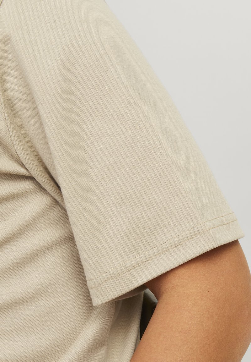 Chemise beige à manches courtes avec un tissu lisse et texturé. L'ourlet est cousu régulièrement, offrant une finition nette au bord des manches.