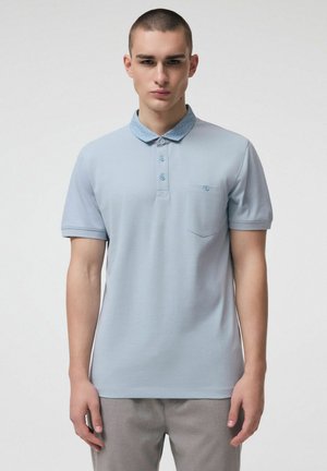 Młody mężczyzna ubrany w jasnoniebieską koszulkę polo z krótkim rękawem, z fakturowanym kołnierzykiem i przednią kieszenią, stojący na jednolitym tle.