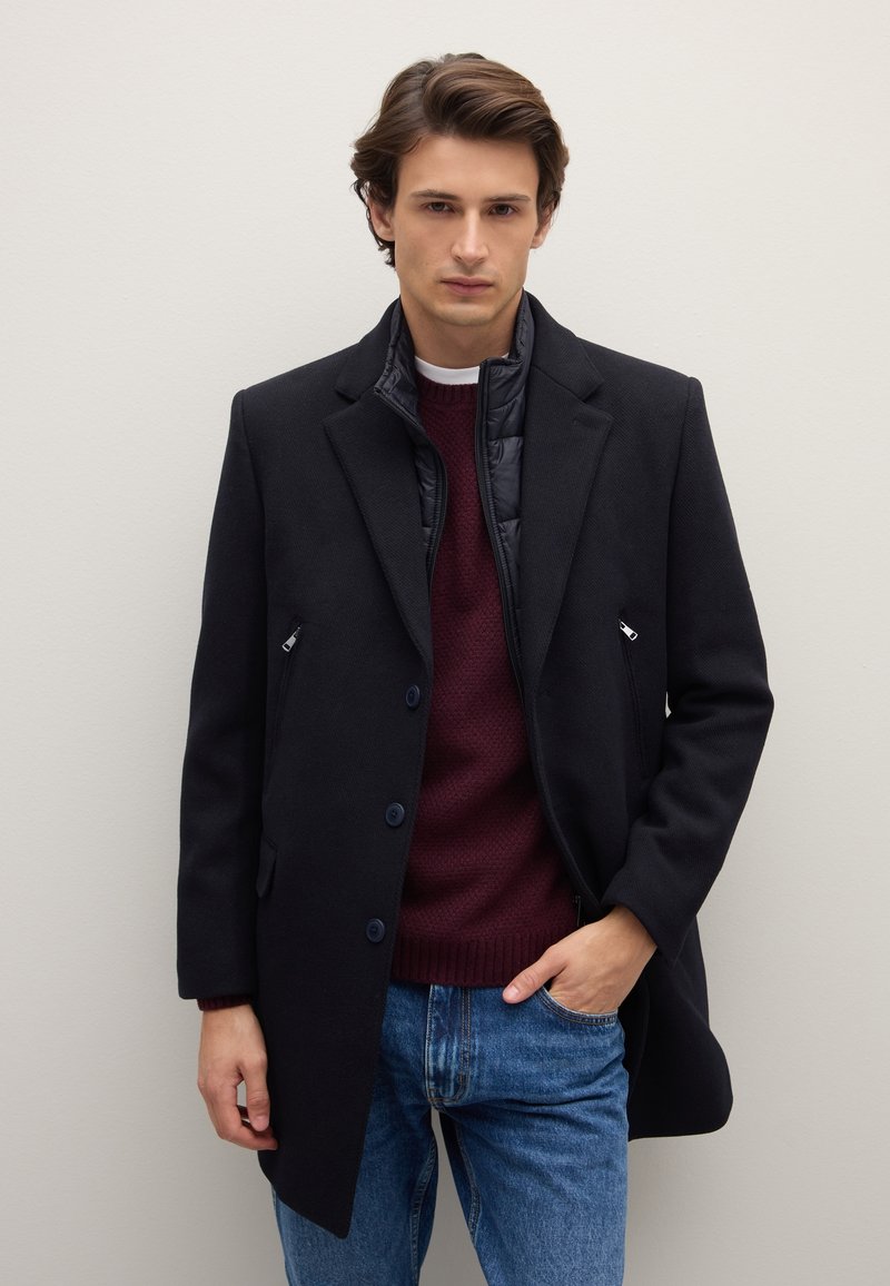 Cappotto blu navy in misto lana con vestibilità sagomata, caratterizzato da un davanti strutturato, due bottoni e tasche laterali; indossato sopra un maglione lavorato a maglia color marrone scuro e jeans.