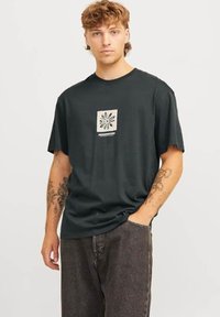 Camiseta de algodón de color verde oscuro con mangas cortas, que presenta un diseño gráfico central en beige de una flor. El modelo la combina con pantalones de mezclilla grises.