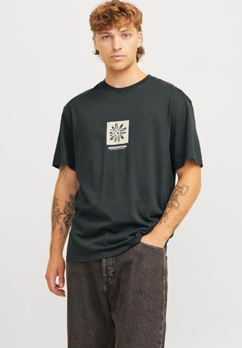 Camiseta de algodón de color verde oscuro con mangas cortas, que presenta un diseño gráfico central en beige de una flor. El modelo la combina con pantalones de mezclilla grises.