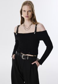 Zwarte off-shoulder gebreide trui met lange mouwen, voorzien van leren bandjes met zilveren accenten. Gecombineerd met hooghangende zwarte broeken en een decoratieve gesp.