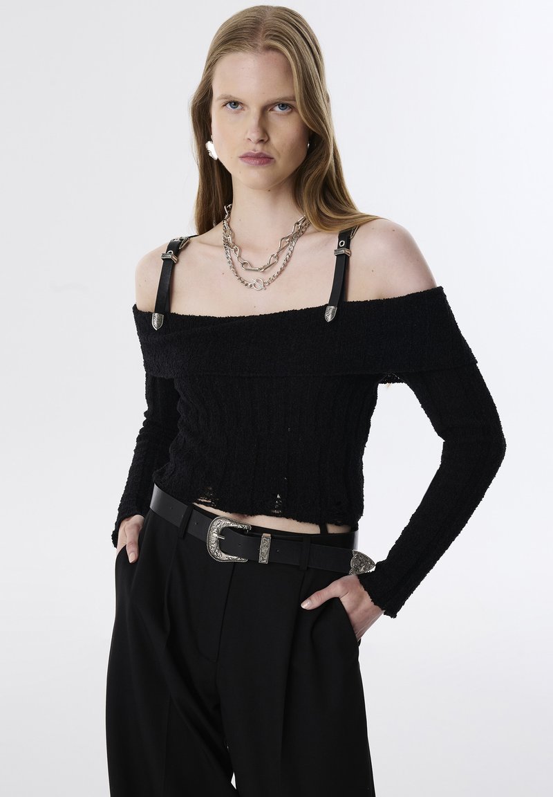 Zwarte off-shoulder gebreide trui met lange mouwen, voorzien van leren bandjes met zilveren accenten. Gecombineerd met hooghangende zwarte broeken en een decoratieve gesp.