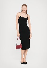 Black midi dress with thin gold chain straps, form-fitting design, smooth texture. Paired with a small red handbag and black pointed shoes.