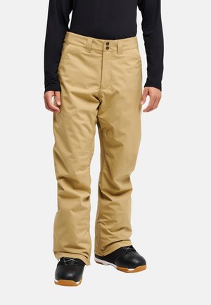 Person trägt beige Schneehose und schwarze Schneestiefel mit braunen Sohlen, von der Taille bis zu den Füßen vor weißem Hintergrund dargestellt.